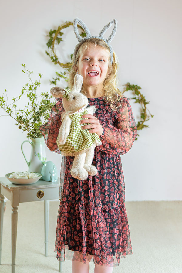 Roześmiana dziewczynka w sukience z opaską z uszami królika i króliczkiem zabawką w rękach na tle wiosennych dekoracji do sesji przedszkolnej.