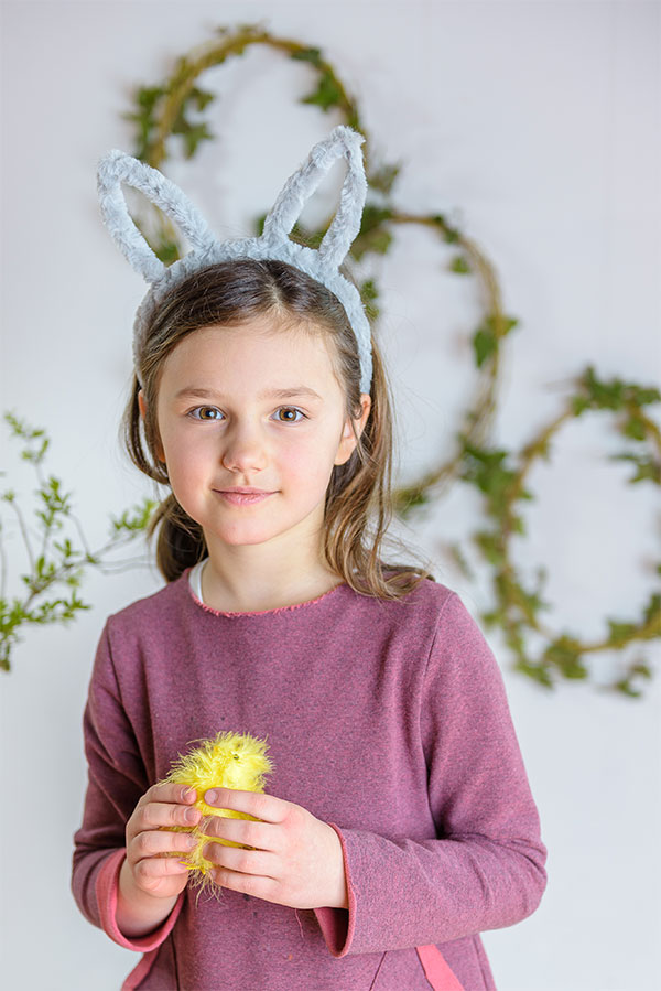 Uśmiechnięta dziewczynka z kurczakiem i opaską na włosach z uszami królika na tle zielonych wieńców świątecznej dekoracji.